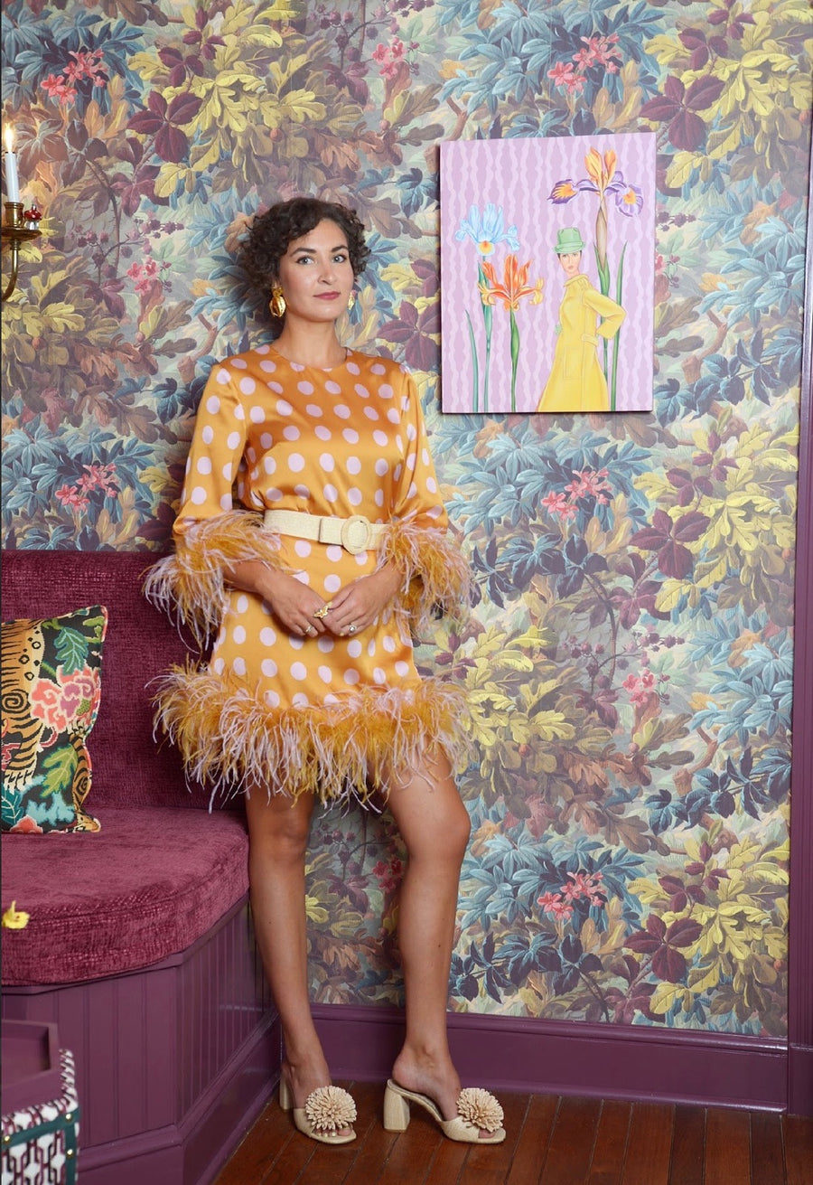 Ashton standing in a feathered gold dress beside one of her paintings