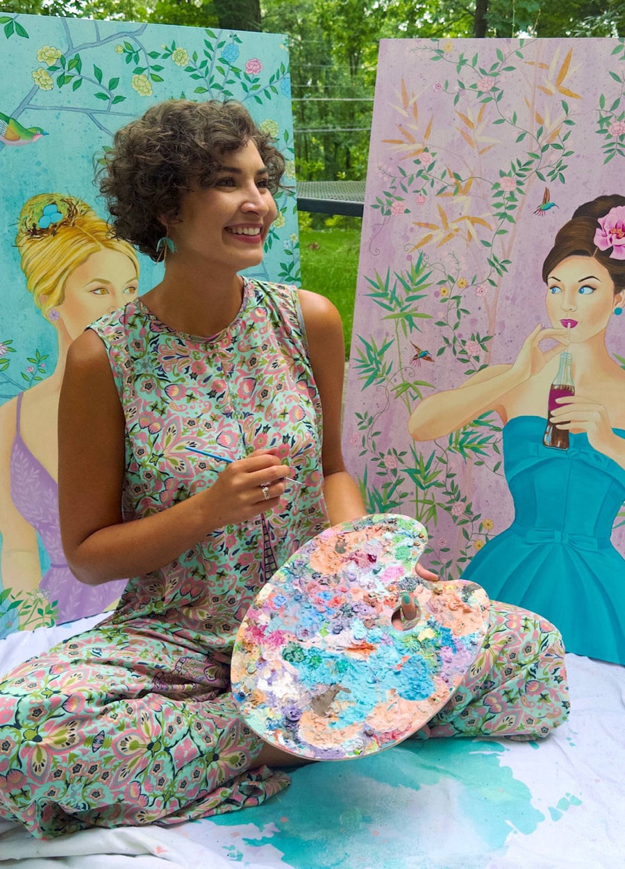 Ashton sitting outdoors with her palette between two of her portraits