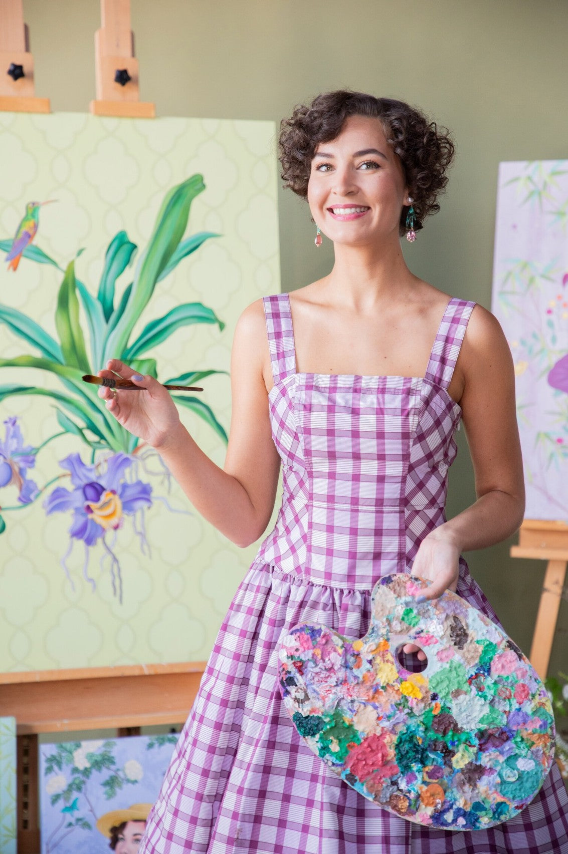 Ashton in a gingham dress holding her palette in front of her paintings